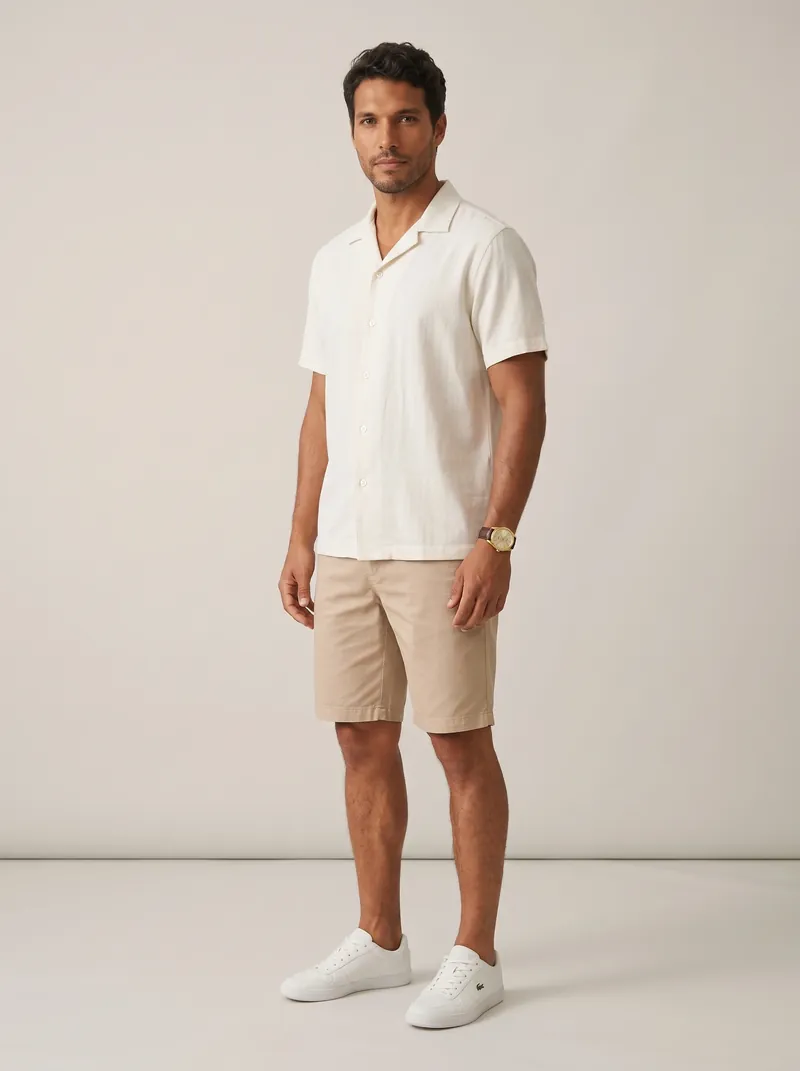 Homem em estúdio claro com camisa gola cubana off-white de manga curta, bermuda de sarja bege, tênis branco de cano baixo e relógio dourado com pulseira marrom