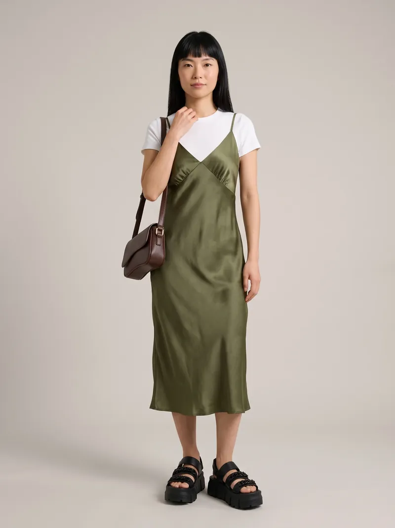 Mulher de franja e cabelo preto liso usa slip dress midi de cetim verde-oliva sobre baby tee branca, bolsa marrom no ombro e sandália flatform preta, em estúdio