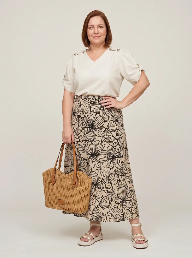 Modelo plus size em estúdio com saia midi evasê estampada, blusa off-white com botões nos ombros, flatform com tachas e bolsa trançada na mão
