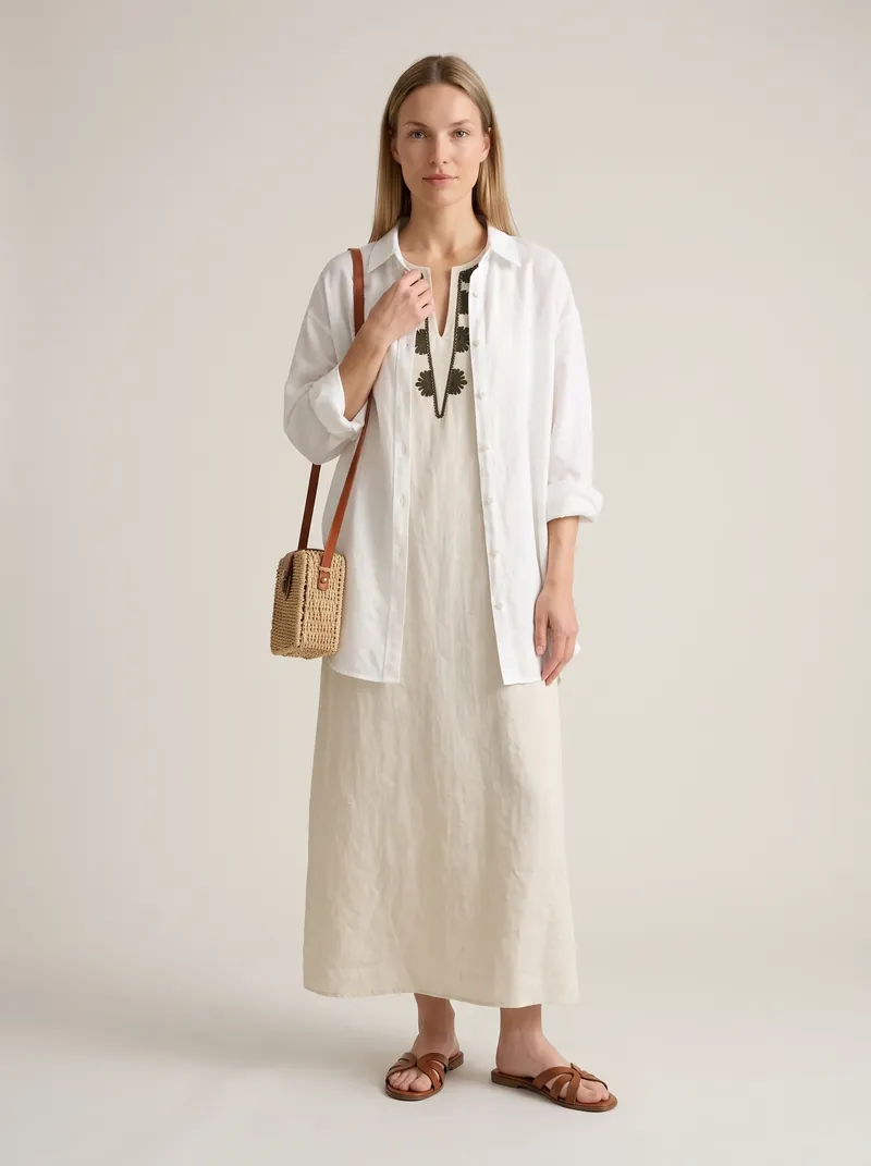 Mulher loira de vestido longo de linho cru com bordado verde no decote, camisa branca oversized aberta, rasteira caramelo e bolsa de palha transversal, em estúdio claro